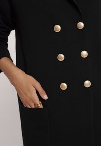 Czarny wełniany płaszcz z podwójnym zapięciem i złotymi guzikami, z bocznymi kieszeniami i prążkowaną fakturą. Dłoń z pierścionkami w kieszeni.