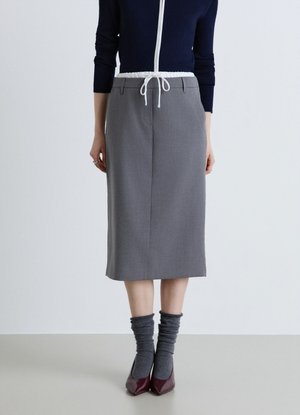 Donna che indossa una gonna grigia al ginocchio con vita con coulisse bianca, calze scure, tacchi bordeaux e una maglia a maniche lunghe blu navy con cerniera bianca.