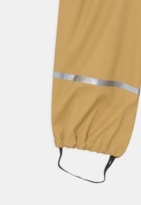 Poncho de chuva na cor mel com bainha ajustada, faixa refletora prateada e detalhe do logótipo. Feito de material liso e impermeável.