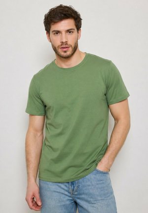 Jeune homme aux cheveux courts et bruns portant un t-shirt vert uni à manches courtes et un jean bleu clair, debout devant un fond clair uni.