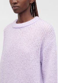 Femme aux cheveux tressés portant un pull en maille doux, violet clair et duveteux avec un col rond sur un fond uni.