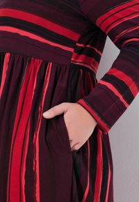Bordeauxrotes Kleid mit vertikalen Streifen in Rot und Schwarz. Es hat einen runden Ausschnitt, lange Ärmel und eine Seitentasche im Rock.