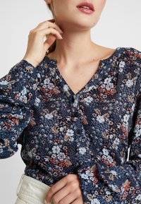 Bluse mit Blumenmuster auf dunkelblauem Hintergrund, die kleine rote und hellblaue Blumen zeigt, lange Ärmel, einen V-Ausschnitt und Knopfdetails hat.