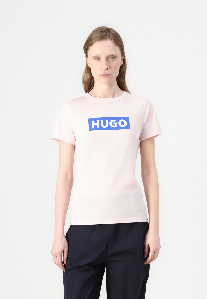 Lys rosa t-skjorte i bomull med en iøynefallende blå rektangulær "HUGO" logo foran. Korte ermer og klassisk rund hals-design.