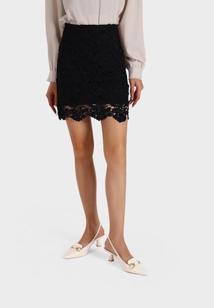 Mini gonna in pizzo nero con motivi floreali e orlo scallop, abbinata a scarpe slingback color beige pallido con un accento dorato.