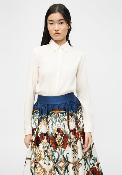 Femme aux longs cheveux noirs portant un chemisier blanc boutonné et une jupe à motifs avec des fleurs rouges ainsi que des dessins bleus, dorés et verts.