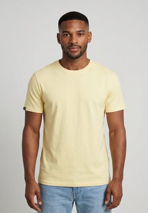 Mann mit kurzen Haaren und Bart, der ein hellgelbes kurzärmeliges T-Shirt und hellblaue Jeans trägt, vor einem einfarbigen grauen Hintergrund.