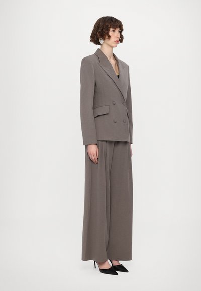 Mittelgrauer doppelt geknöpfter Blazer mit breiten Revers, kombiniert mit passenden weiten Hosen und schwarzen spitzen High Heels. Glatte Textur.