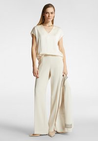 Top in raso crema con scollatura a V e maniche a cappuccio, abbinato a pantaloni larghi crema. Il modello tiene in mano un giubbotto coordinato decorato con perline.