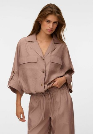 Femme portant une chemise ample taupe à boutons avec poches à rabat et un pantalon assorti à cordon, debout avec une main dans la poche sur fond blanc.