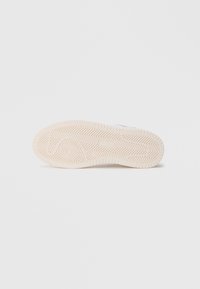 Zapatilla blanca con una suela de goma texturizada que presenta un patrón de espiga y un detalle circular en el talón. Material superior liso.