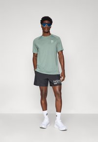 Ανοιχτό πράσινο αθλητικό μπλουζάκι με λογότυπο, μαύρο σορτσάκι με λευκό κείμενο Nike, λευκά αθλητικά παπούτσια και μπλε γυαλιά ηλίου. Λείος υφασμάτινος σχεδιασμός, άνετη εφαρμογή.