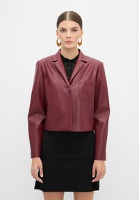 Giacca cropped in ecopelle bordeaux con chiusura a un bottone, rever a punta e taschino sul petto, indossata sopra un top in pizzo nero.