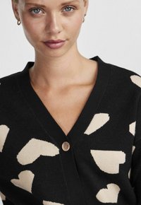 Cardigan noir avec grands motifs de cœurs beiges, col en V, et une fermeture à un bouton sur le devant. Texture douce.