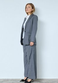 Traje gris con un sutil patrón de puntos, americana de un solo botón y pantalones de pierna ancha. Combinado con una camisa blanca y zapatos de tacón negros.