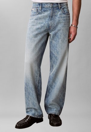 Jasnoniebieskie jeansy o prostych nogawkach z delikatnym kwiatowym wzorem, noszone z czarnymi mokasynami i srebrną bransoletką na prawej ręce.