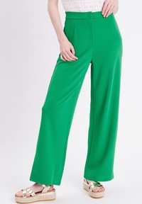 Femme portant un pantalon ample taille haute vert fluo avec un bouton, un haut blanc et des sandales à plateforme métalliques sur fond blanc.