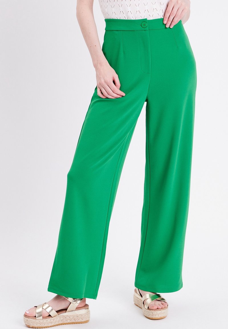 Femme portant un pantalon ample taille haute vert fluo avec un bouton, un haut blanc et des sandales à plateforme métalliques sur fond blanc.