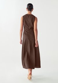 Vestido maxi marrón sin mangas con un torso ajustado, falda acampanada, textura sutil y cierre de cremallera en la parte trasera. Llevado con tacones de color claro.