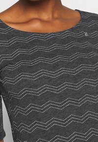 Top de tela gris con un escote ancho y mangas tres cuartos, adornado con un patrón de zigzag blanco y pequeños acentos de puntos.