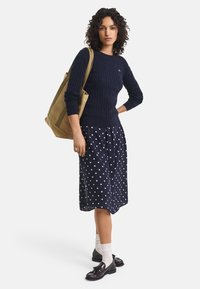 Maglione blu a trecce, gonna a pois e scarpe nere. La modella porta una borsa a tracolla color cammello. Tessuto misto cotone, design casual.