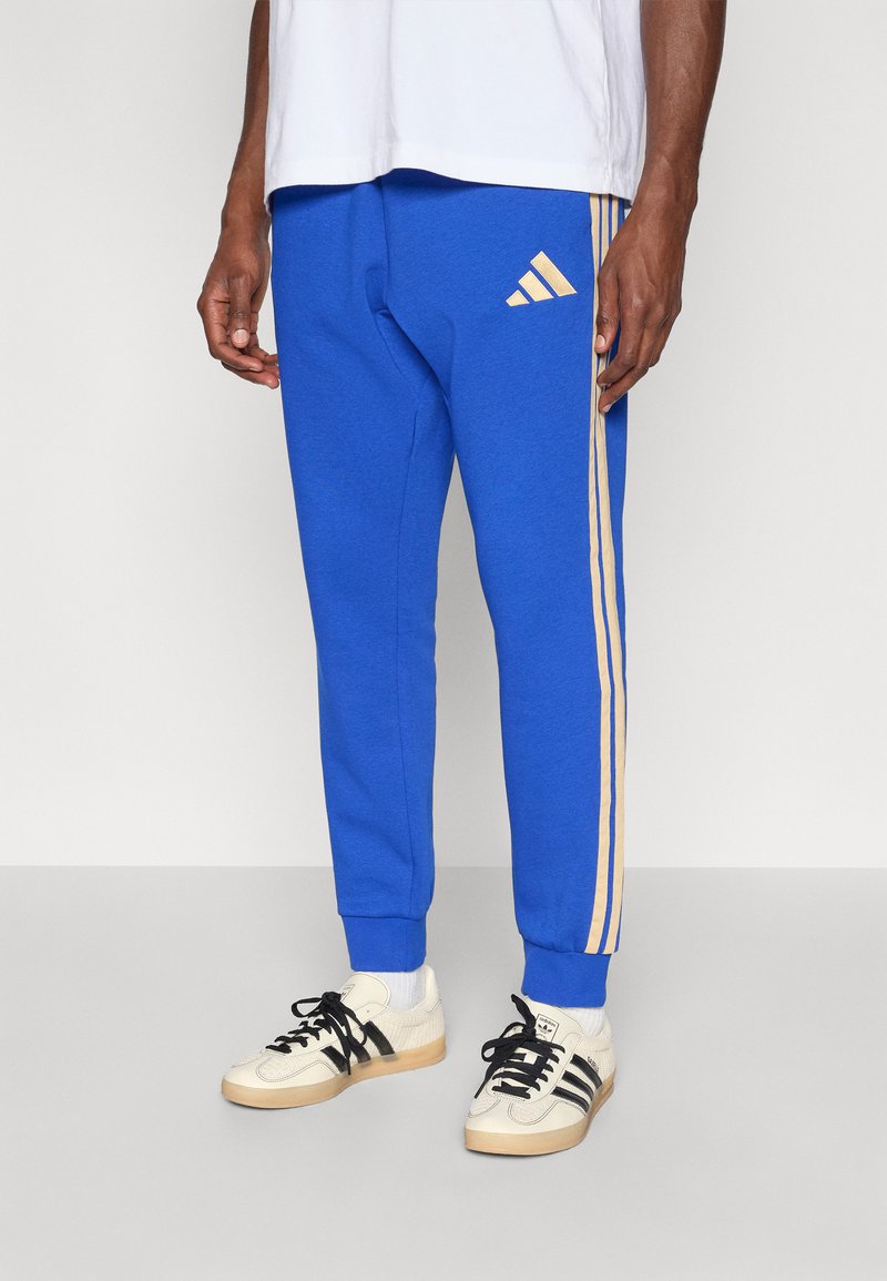 Modré teplákové nohavice z mäkkého materiálu, s dvoma zlatými pruhmi po bokoch a logom Adidas na ľavom boku. V kombinácii s bielymi topánkami.