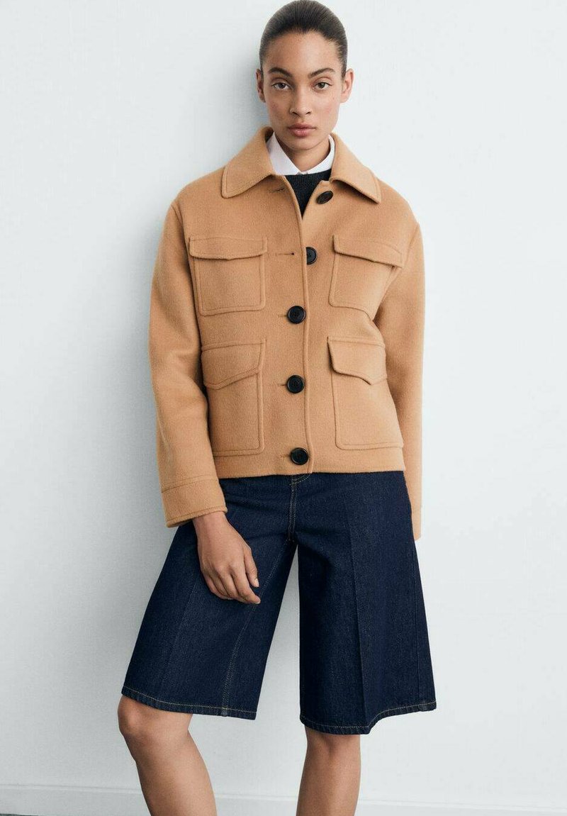 Vrouw in een camelachtige jas met knopen en vier klepzakken, gedragen over een zwarte top en donkerblauwe denim short tot de knieën.
