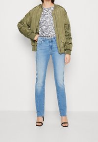 Veste bomber vert olive associée à un t-shirt gris à motif léopard, un jean slim bleu clair et des sandales à talons hauts noires.