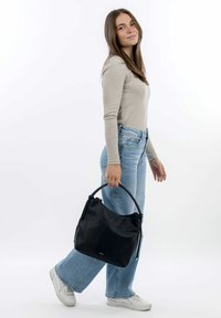 Borsa hobo in pelle nera con superficie texturizzata, chiusura con zip e manico rotondo, sostenuta da una persona che indossa jeans azzurri e sneakers bianche.
