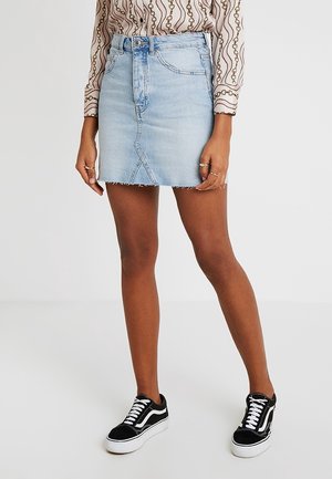 Person wearing light blue denim mini skirt with frayed hem, patterned long-sleeve blouse, and black and white sneakers standing on plain background.