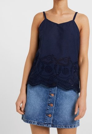 Kobieta w granatowej, bezrękawnikowej bluzce z haftowanym dołem i dżinsowej mini spódniczce zapinanej na guziki, stojąca na jednolitym tle.