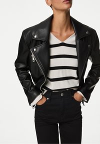 Chaqueta biker de cuero negro con cremalleras plateadas, llevada sobre un suéter de rayas blancas y negras con escote en V, combinada con jeans negros.