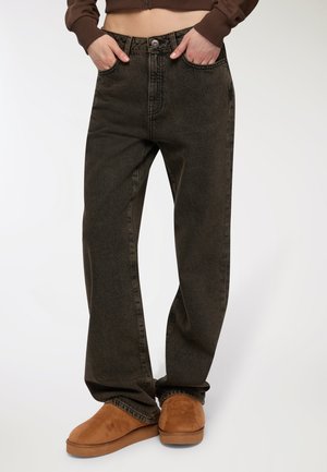 Jeans a gamba larga di colore verde scuro realizzati in denim, con chiusura a bottone, passanti per cintura e tasche frontali, abbinati a scarpe slip-on marroni.