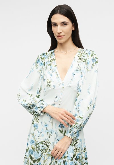 Femeie cu păr lung și închis la culoare poartă o rochie albă cu imprimeu floral, cu mâneci lungi bufante și un decolteu adânc în V, stând în fața unui fundal simplu.