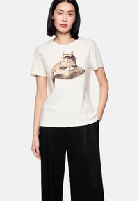 Baumwoll-T-Shirt in Off-White mit einer Katzenabbildung, die eine Brille trägt, kurze Ärmel und Rundhalsausschnitt, kombiniert mit lockeren schwarzen Hosen.
