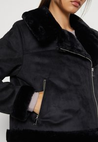 Svart jacka med fuskpälsfoder, med dragkedja, sidofickor och bred krage. Kroppen har ett mjukt, texturerat tyg för värme.
