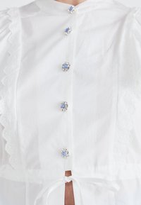 Hvit bluse med puffede ermer, blonder, runde blå og sølvknapper, og knyttet midjedetalj. Glatt stoffstruktur.