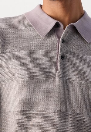 Mężczyzna w teksturowanej dzianinowej koszulce polo w jasnoszarym kolorze z fioletowym kołnierzykiem i dwoma czarnymi guzikami przy szyi.