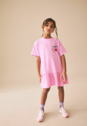 Jeune fille portant une robe rose avec un motif de cerises, des chaussettes violettes et des baskets roses, debout devant un fond beige uni.