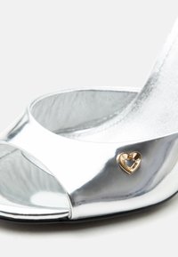 Ασημένια σανδάλι με ψηλό τακούνι, ανοιχτό δάχτυλο και μικρή χρυσή καρδιά ως διακοσμητικό στο μπροστινό λουράκι.