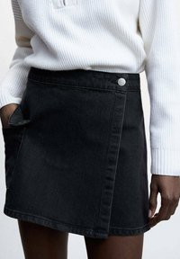Minifalda de mezclilla negra con diseño envolvente, acento de botón plateado y bolsillo lateral. La textura es suave con detalles de costura visibles.