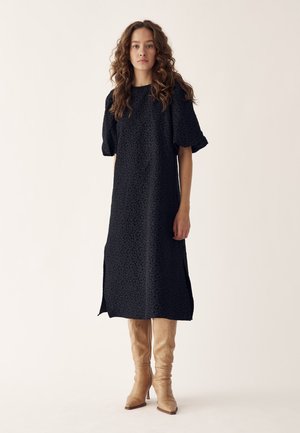 Sieviete ar cirtainiem matiem, tērpusies melnā midi kleitā ar uzpampušām piedurknēm un šķēlumiem, valkājot bēšas ceļgalu augstas zābaki, stāv pret vienkāršu fonu.