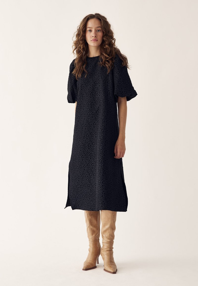 Femeie cu păr creț purtând o rochie midi neagră cu manșete bufante și fante, asortată cu cizme bej până la genunchi, stând în fața unui fundal simplu.