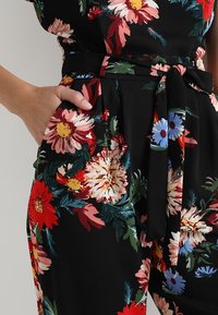 Combinaison florale avec une base noire, de grandes fleurs rouges et roses, un feuillage vert, et une ceinture assortie nouée en un nœud à la taille.