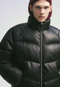 Veste matelassée en cuir noir avec un col montant, fermeture zippée, texture matelassée et léger éclat. Fini lisse avec des détails aux poignets.
