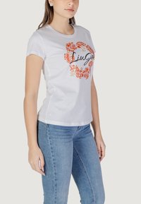 Weißes Baumwoll-T-Shirt mit einem orangefarbenen Blumendesign und einem schwarzen Schriftzug-Logo. Kurze Ärmel und eine figurbetonte Passform, kombiniert mit blauen Jeans.