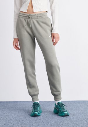 Personne portant un pantalon de survêtement gris Nike, des baskets Nike turquoise, des chaussettes blanches, et un top blanc court en côte à manches longues, sur fond uni.