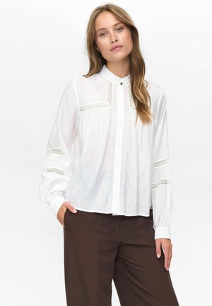 Femme portant un chemisier blanc à manches longues avec des détails en dentelle et un pantalon marron, se tenant avec une main dans la poche devant un fond uni.