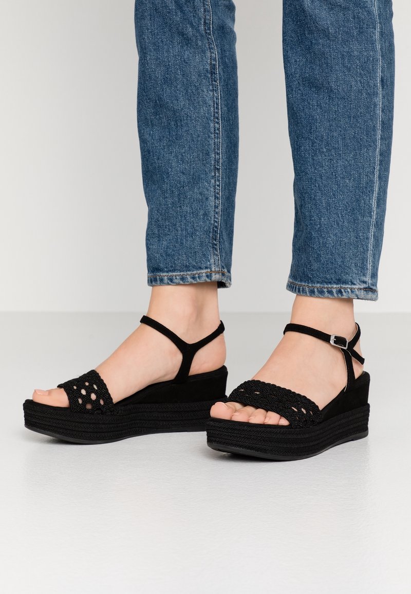 Pieds portant des sandales à plateforme noires avec des brides tressées et une boucle à la cheville, associées à un jean bleu.