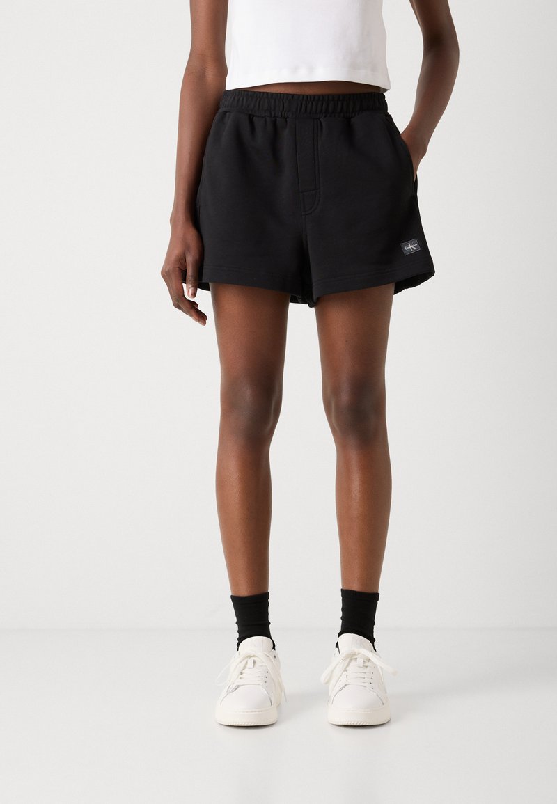 Person iført sorte shorts med elastisk talje med Calvin Klein-logo, hvide sneakers, sorte sokker og en hvid ærmeløs top mod en ensfarvet baggrund.